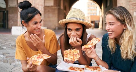Three beautiful women sitting on the stairs of the city streets eating pizza from a street stall. The happy girls enjoy the week
