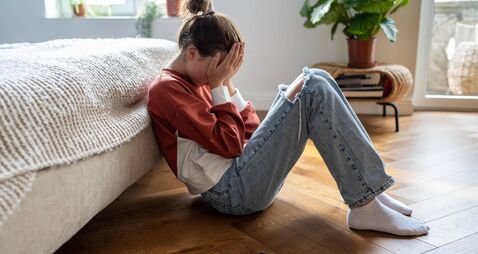 Unhappy teen girl covering face with hands crying while sitting on floor with mobile phone nearby